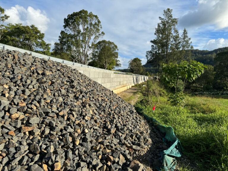 A pile of large rocks is in the foreground, next to a retaining wall. Trees and a grassy area are seen to the right, with a cloudy sky above. Hills in the background suggest a rural or semi-rural location.