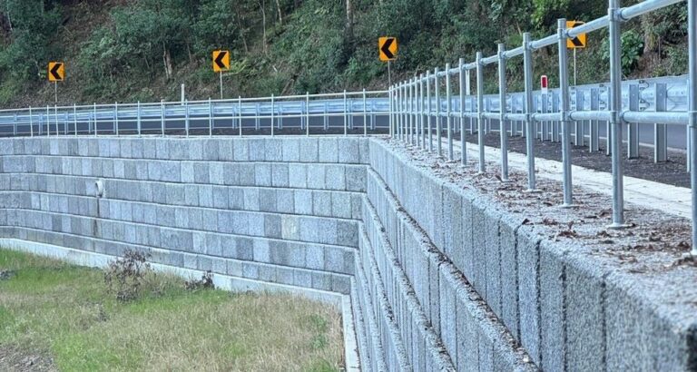 A concrete retaining wall supports a road lined with metal railings. Yellow directional signs indicate a curve ahead. Dense green foliage is visible in the background.