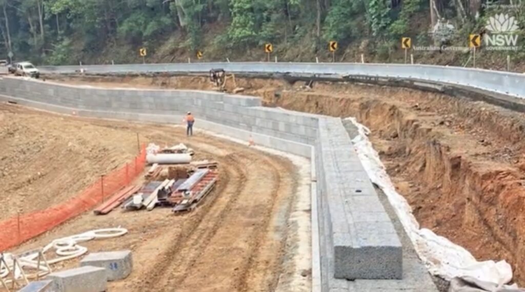 A construction site with a newly built retaining wall along a curved road. Piles of construction materials are visible, and a worker in an orange vest is on site. Trees line the background, and safety barriers parallel the road.